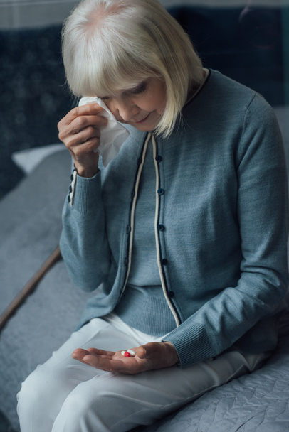 smutny starszy kobieta siedzi na łóżku, trzymając pigułki i ocierając łzy z chusteczką w domu - Zdjęcie, obraz