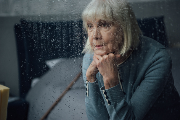mujer mayor molesta sentada y apoyando la barbilla con las manos en casa a través de la ventana con gotas de lluvia
 - Foto, Imagen