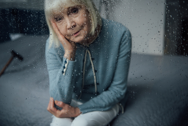eenzame senior vrouw zitten en kijken naar camera door raam met regendruppels - Foto, afbeelding