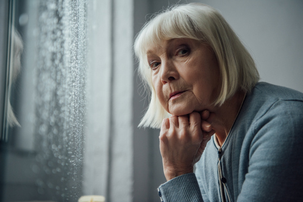 pensativa mujer mayor con el pelo gris apoyando la barbilla con la mano y mirando a la cámara en casa
 - Foto, Imagen