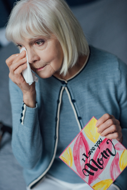 senior woman holding card with 'i love you mom' lettering, crying and wiping tears with handkerchief at home - Photo, Image