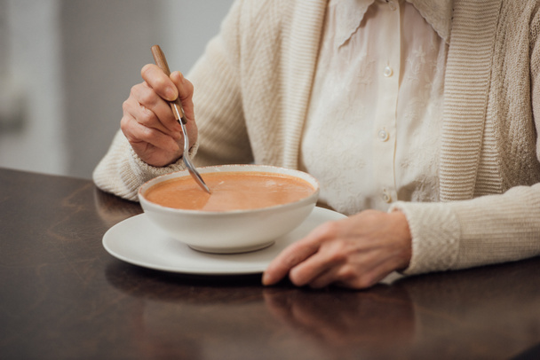 частичный вид пожилой женщины, сидящей за столом и кушающей сливочный суп дома
 - Фото, изображение