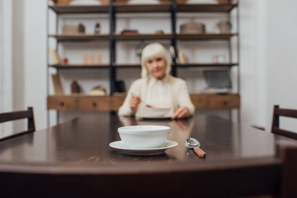селективный фокус миски и ложки на столе со старшей женщиной на заднем плане
 - Фото, изображение