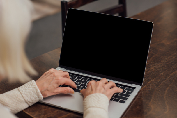 bijgesneden weergave van senior vrouw zittend aan tafel en typen op laptop met leeg scherm thuis - Foto, afbeelding