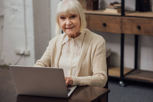 usměvavá starší žena sedí u stolu a psaní na notebooku doma - Fotografie, Obrázek