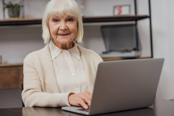 smiling senior woman sitting at table, looking at camera and typing on laptop at home - Foto, imagen