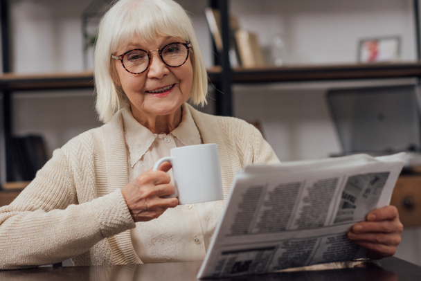 щаслива старша жінка сидить за столом і читає газету під час пиття кави вдома
 - Фото, зображення