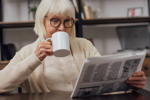 Senior vrouw in glazen aan tafel zit en krant lezen terwijl het drinken van koffie thuis - Foto, afbeelding