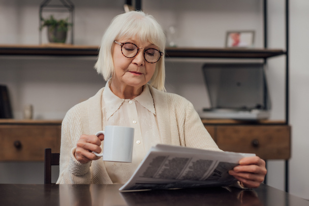 Seniorin in Gläsern sitzt am Tisch, trinkt Kaffee und liest Zeitung zu Hause  - Foto, Bild