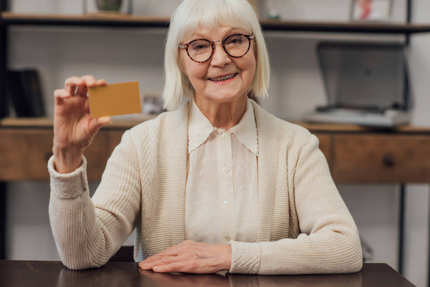 masada oturan, kredi kartı evde tutan ve kameraya bakarak mutlu kadın kıdemli  - Fotoğraf, Görsel