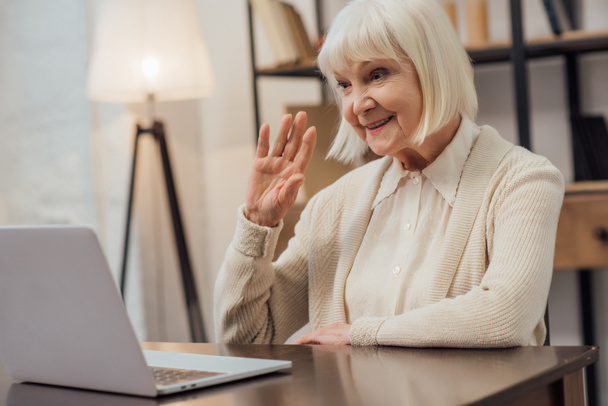 усміхнена старша жінка сидить за комп'ютерним столом і махає під час відеодзвінка вдома
 - Фото, зображення