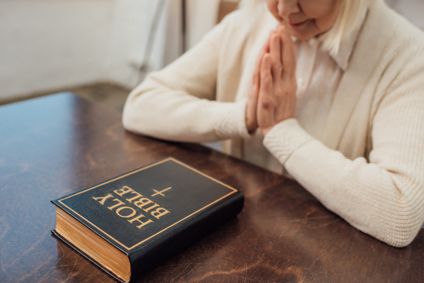 visão parcial da mulher idosa sentada e orando na frente da Bíblia sagrada em casa
 - Foto, Imagem