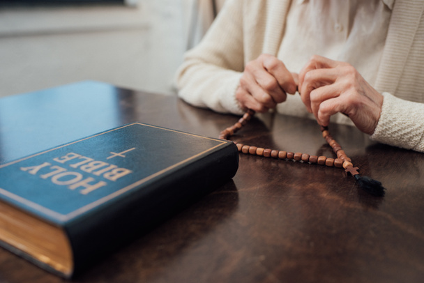 partial view of senior woman sitting at table with rosary and holy bible at home - Foto, afbeelding