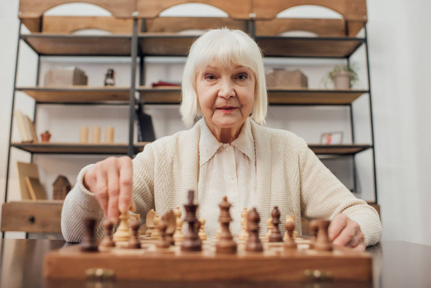 senior woman sitting at table, looking at camera and playing chess at home - Fotoğraf, Görsel
