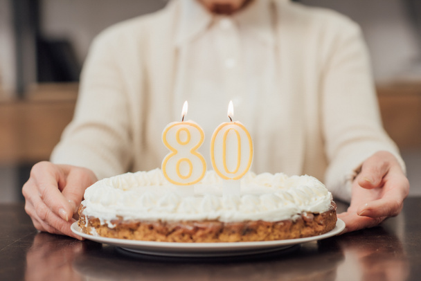 partial view of senior woman holding  birthday cake with number 80 on top at home  - Foto, Imagem