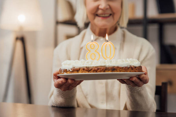 cropped view of smiling senior woman holding birthday cake with number 80 on top at home  - Foto, afbeelding