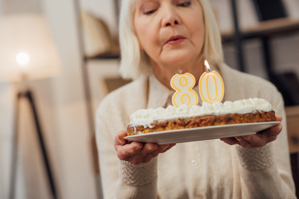 年配の女性が自宅の誕生日を祝っている間番号 80 の上でケーキの上のろうそくを吹き  - 写真・画像