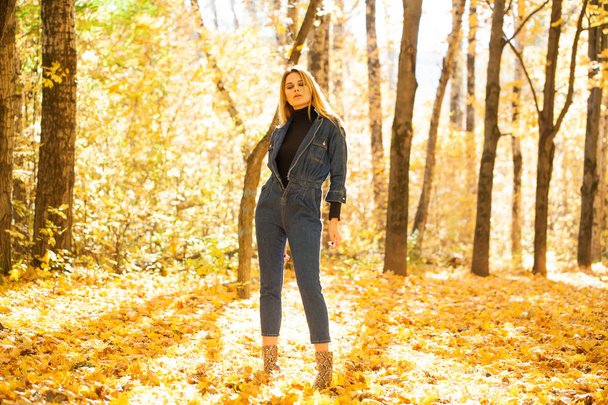 Full portrait of Stylish young blonde woman in blue denim overalls in autumn park - Фото, зображення