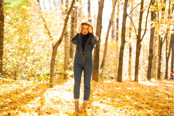 Full portrait of Stylish young blonde woman in blue denim overalls in autumn park - Zdjęcie, obraz