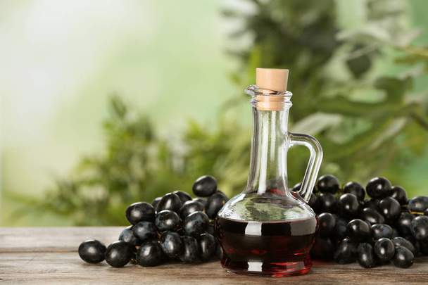 Glass jug with wine vinegar and fresh grapes on wooden table against blurred background - Foto, Bild