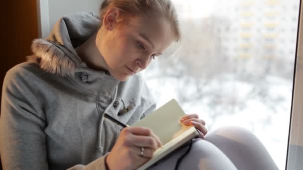 Chica joven escribiendo en su diario mientras está sentada en una ventana grande
 - Metraje, vídeo