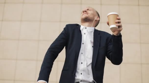 Attractive young businessman listening to music in headphones on smartphone, standing outside the office and funny dancing to the rhythm. Drink coffee, funny dancing moves. Slow motion - Séquence, vidéo