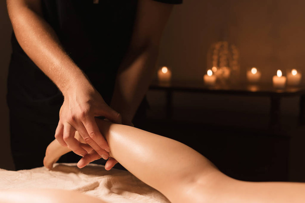 Close-up of male hands doing calf massage of female legs in a dark room with candles in the background. Cosmetology and spa treatments. - Foto, afbeelding