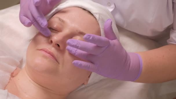 美容師は、中年女性の顔の目の周りのクリームを入れて薄紫色の手袋で手します。美容室美容クリニックで治療上のプロシージャ。クローズ アップ. - 映像、動画