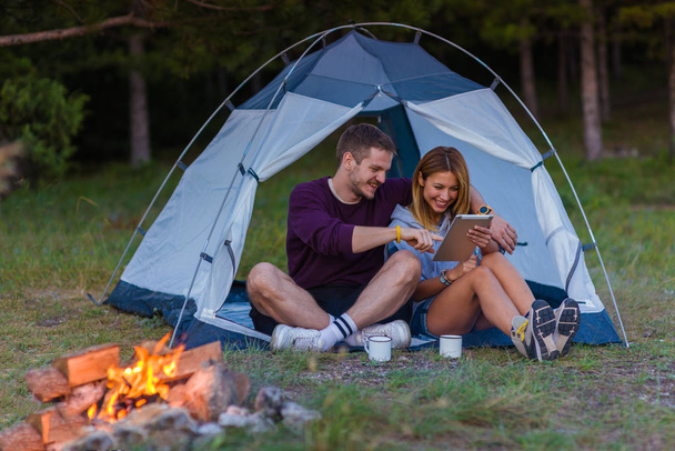 若いカップル コーヒーを飲みながら、山の景色を楽しんで、日没のたき火でタブレットをチェック - 写真・画像