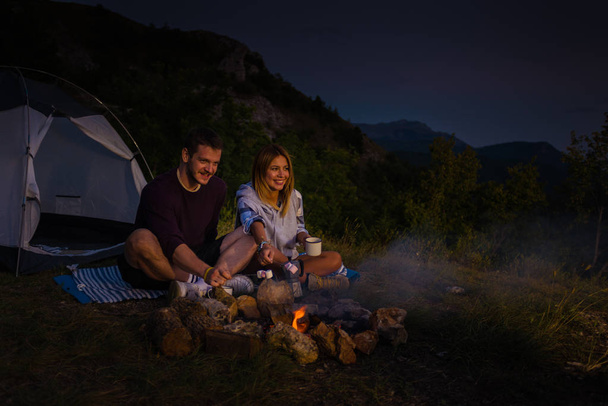 Young couple relaxing by the campfire, eating rosted marshmallows and drinking coffee in the forest hill in the dusk - Photo, Image