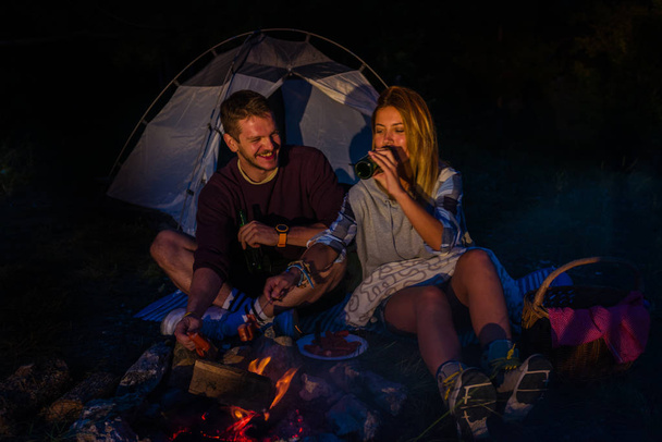 Man and woman rosting sausages on the campfire, drinking beer and relaxing by the campfire in the mountain top in the night. Romantic campfire concept. - Photo, Image