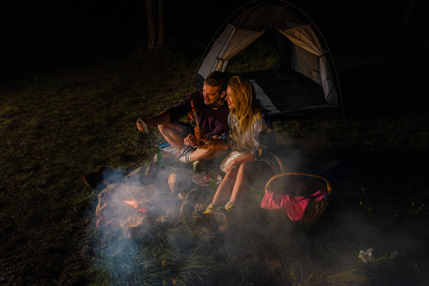 Jong koppel selfie maken, bakken op het kampvuur worstjes en bier drinken in de heuvel bos in de nacht. Ontspannen romantische uitje weekend - Foto, afbeelding