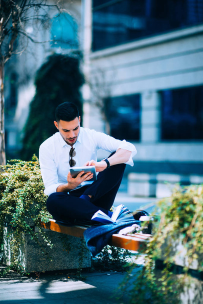 ein gutaussehender Mann in weißem Hemd sitzt auf der Bank und schaut in sein weißes Tablet. - Foto, Bild