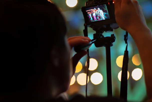 Uomo movimento offuscata utilizzando la fotocamera con treppiede con luci scintillanti bokeh in background. Ritratto candido da dietro dell'uomo che scatta la foto
 - Foto, immagini