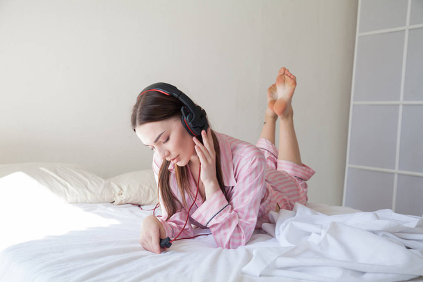 ピンクのパジャマを着てベッドの上でダンスのヘッドフォンで音楽を聴く女性 - 写真・画像