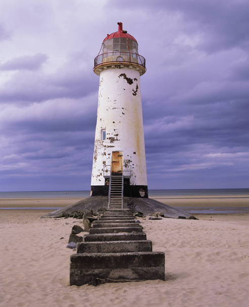 An old white light house with a bright red roof sitting on a sandy beach with a stone pathway infront, the sea is in the distance and a big blue sky with white swirling clouds is behind the light house, Talacre lighthouse, Prestatyn, Wales, United Ki - Photo, Image