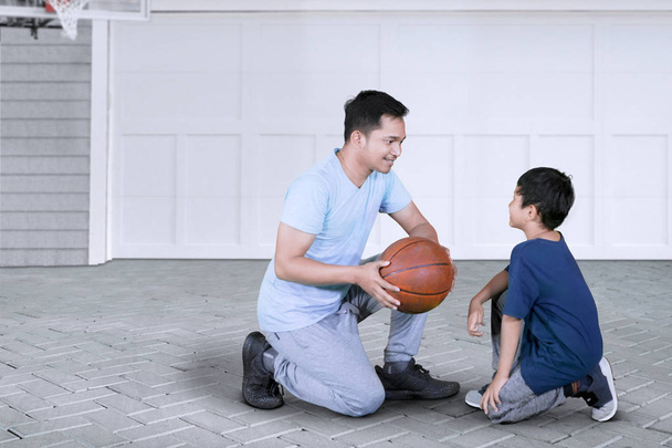 Picture of young man kneeling in the garage while teaching his son to play basketball - Zdjęcie, obraz