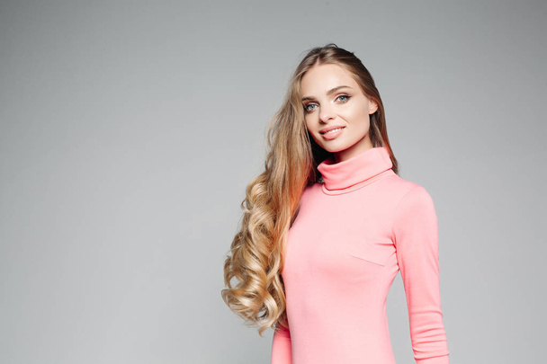 Studioporträt einer schönen Geschäftsfrau blond mit blauen Augen und langen Haaren, trägt ein elegantes rosafarbenes Kleid und hält ihre Hand auf der Taille. isoliert auf grauem Hintergrund - Foto, Bild