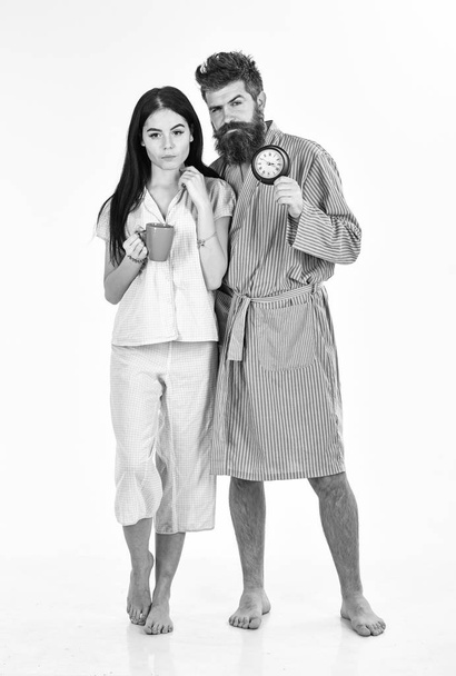 Perfect morning concept. Couple, family woke up on time. Couple in love, young family in pajama, bathrobe stand isolated on white background. Girl with coffee cup, man holds clock in hand - Φωτογραφία, εικόνα