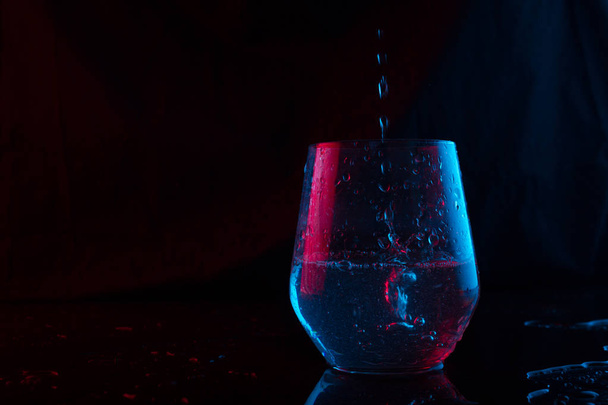 Water highlighted in blue and red splashing into a glass goblet on a black background - Foto, Bild
