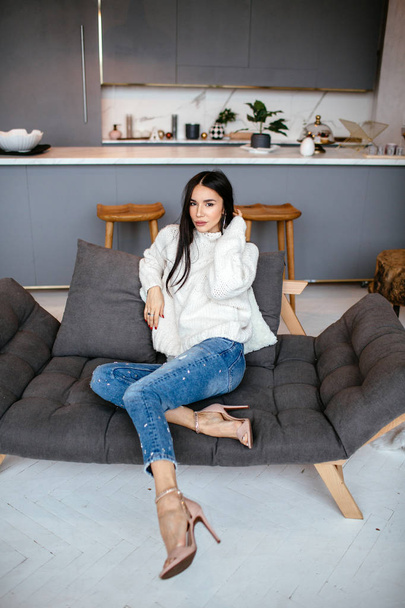 Menina bonita em uma jaqueta branca e jeans está descansando no sofá em casa. retrato, estilo escandinavo
 - Foto, Imagem