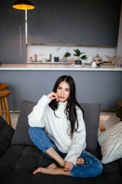 Beautiful girl in a white jacket and jeans is resting on the sofa at home. portrait, scandinavian style - Fotó, kép