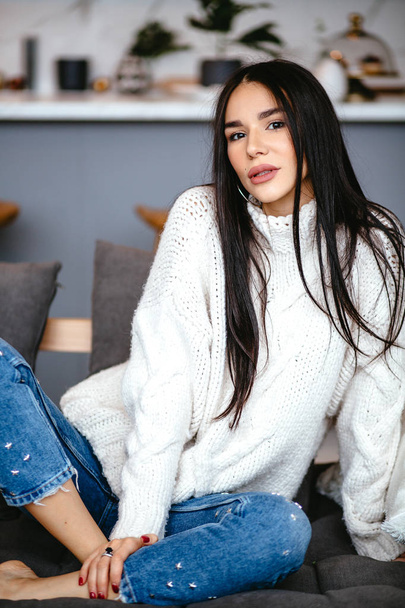 Beautiful girl in a white jacket and jeans is resting on the sofa at home. portrait, scandinavian style - Photo, Image
