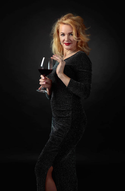Atractiva mujer de mediana edad en vestido de noche negro con copa de vino tinto. Feliz cuarenta años rubia sobre un fondo negro
. - Foto, imagen