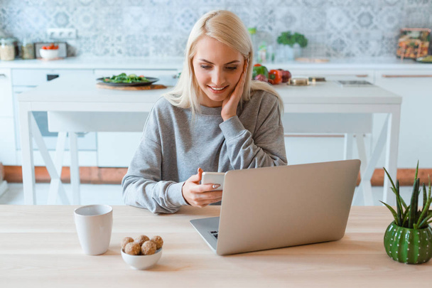 Surprised blonde young woman sitting at the table in the kitchen and using smart phone. Laptop on the table.  - Фото, изображение
