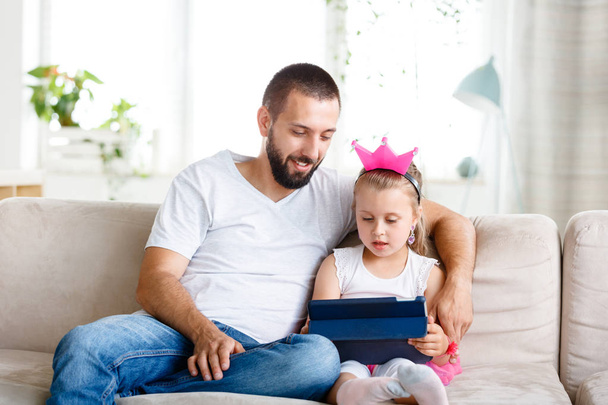 Father and daughter sitting on sofa at home and using digital tablet together.  - Foto, afbeelding
