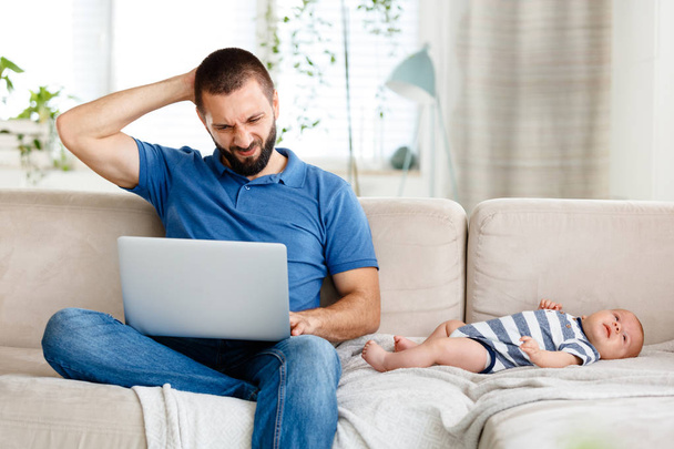 Worried young father working from home, using laptop while his baby boy lying on sofa next to him.  - Foto, immagini