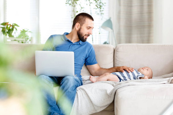 Young man sitting on sofa at home and using laptop while his baby boy lying next to him.  - 写真・画像