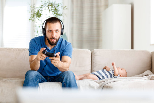 Excited young man sitting on sofa at home and playing video game while his baby boy lying next to him.  - Foto, afbeelding
