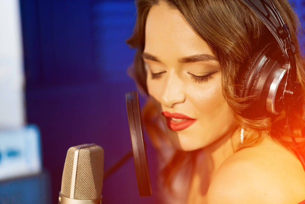 Caucasian female singer with headphones and closed eyes sings at the mic in a recording studio. Portrait of a beautiful woman in headphones singing. Closeup photo. - Fotografie, Obrázek